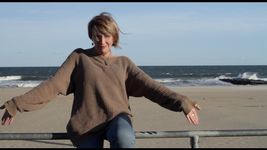 Kate Bornstein stands by a railing at the beach on a sunny day, facing the camera with their arms stretched out to their sides catching the breeze. She is a white person with a blonde short bob hairstyle. She is wearing a loose oversized sweater and jeans.