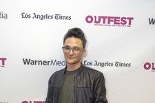 Sam Feder posing on a red carpet in front of a white backdrop with festival sponsor logos. Sam is a white person with short hair and glasses. They are wearing a leather motorcycle jacket on top of a dark shirt.