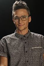 Sam Feder captured with a slight smile, seated against a dark background in a professional studio setting. They are wearing a black and white patterned button up shirt with short rolled sleeves.