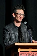 Sam Feder smiling while standing at a podium on stage. Sam is a white person with dark coiffed hair and glasses. They are wearing a leather jacket over a dark shirt.