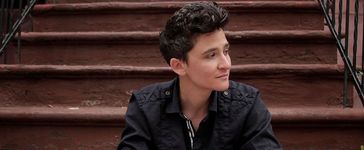 Sam Feder sitting on a brownstone staircase on a street in New York. Sam is a white person with dark coiffed hair and is wearing a dark top. Their face is turned away from the camera.