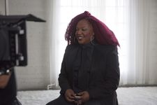 Behind the scenes photograph of Jazzmun during filming on set. Jazzmun is a Black person with purple-red long coily hair. She has silver jewelry and is wearing an all black suit and tie outfit. The set is a spacious and airy white room decorated with sheer curtains and a fluffy white rug.