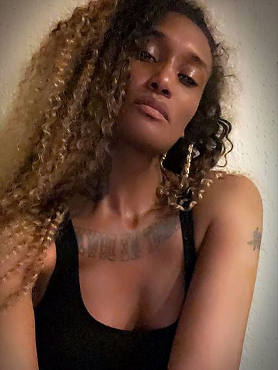 A close cropped portrait of D. Smith. She is a Black woman, with long golden curly hair, and wears a black tank top. The camera angle is positioned upwards, creating the effect that D. Smith is looking slightly down on the viewer.