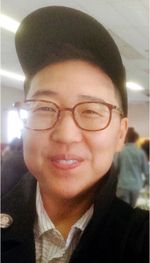 Eric Cho has glasses, a happy grin, and is Korean. They wear a baseball cap and collared shirt under a sweater. The image is closely cropped on thier face but there are people and ceiling tiles in the background which suggests an indoor venue like a school.