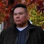 Elwood Jimmy looks at the camera with a neutral expression. Elwood has a short undercut, is Nêhiyaw, and wears a light-weight running jacket. They are outdoors in front of a maple tree tinged red with fall.