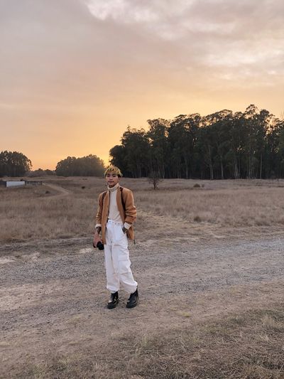 Sal Tran is nestled into a landscape with a road, a field of dried grass, trees, and a sunset sky. Sal Tran is a queer Vietnamese person with edgy short hair. They lazily rest a camera against their thigh and wear a blazer, turtleneck, and cargo pants in a pallet that complements the landscape.