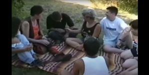 A group of intersex young adults from various backgrounds sits in a circle, cross legged on a blanket in a grassy area. They are dressed in casual summer clothing of the mid 1990s including jean shorts, big t-shirts, vests and tank tops. They are mid conversation with each other.