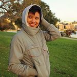 Jaq Nguyen Victor stands with a smile and one hand in their front jacket pocket and the other behind their head. Jaq is a Southeast Asian person with light skin and black hair. They wear a jacket and hoodie in a park.