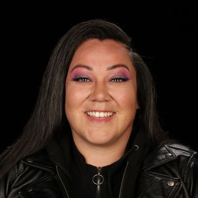 Lacey Hill is an Indigenous person with long black hair, light-skin, and a big smile. She is wearing a black leather jacket and bright pink and purple eyeshadow. She stands in front of a black background and looks directly into the camera.