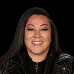 Lacey Hill is an Indigenous person with long black hair, light-skin, and a big smile. She is wearing a black leather jacket and bright pink and purple eyeshadow. She stands in front of a black background and looks directly into the camera.