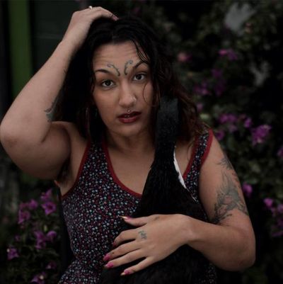 Neve Be(ast) stands with their eyebrows raised and one hand in their hair and the other rested on their chest. Neve Be(ast) is a Black, Arab, and Indigenous mixed race person. They have dark wavy hair to their shoulders, medium-brown skin, a Medusa piercing, curly tattoos above their eyebrows, a few other tattoos on their arms, and pink nails. Behind them is a dark and blurry background of plants with pink flowers.