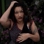 Neve Be(ast) stands with their eyebrows raised and one hand in their hair and the other rested on their chest. Neve Be(ast) is a Black, Arab, and Indigenous mixed race person. They have dark wavy hair to their shoulders, medium-brown skin, a Medusa piercing, curly tattoos above their eyebrows, a few other tattoos on their arms, and pink nails. Behind them is a dark and blurry background of plants with pink flowers.