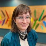 Raphaële Frigon smiles in front of an abstract artwork with bright colours. Raphaële has clear glasses, brown shoulder-length hair with bangs, and light skin. She wears a kercheif around her neck and a satin blue jacket.