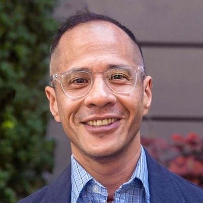 Félix Endara is an Ecuadorian transgender man with light brown skin. He is wearing a navy blue suit with a light blue checkered button up. He has short hair and is wearing clear framed glasses. Félix is in front of a blurred grey wall with a green bush on the left and redish flowers on the right.