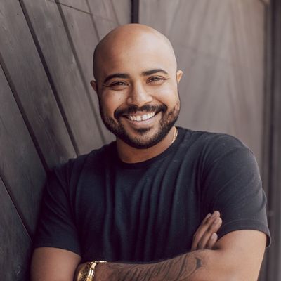 Lucah Rosenberg-Lee is a Black trans man with medium brown skin. He is shown from his chest up with his arms crossed and smiling. Lucah is wearing a short sleeve navy blue t-shirt. He has a tattoo on his right arm and a gold watch. Lucah is bald with dark facial hair and a chain tucked into his t-shirt. Lucah is in front of a dark brown wooden wall.