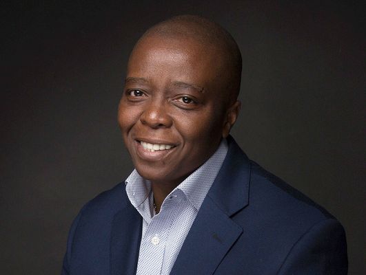Yance Ford is a Black trans man with dark brown skin in front of a black background. He is bald wearing a navy blue suit and a white button up with small light blue dots.
