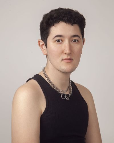 Morgan Sullivan is trans with very light skin in front of a white gray background. They are wearing a black muscle t-shirt with two silver chains shown from the abdomen up. They have short black hair with faded sides.