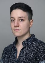 Noah Schamus is trans non binary with light skin with a gray background. They have short black hair with faded sides wearing a short sleeve button up that is navy blue with a busy pattern.