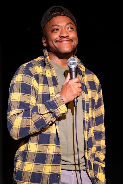 Kai Choyce is a Black trans man with brown skin and black hair. He is seen mid-standup performance, smiling into a microphone in front of a black background. He is wearing jeans, a backwards black baseball cap, and a yellow-and-black plaid sweater over an olive shirt.