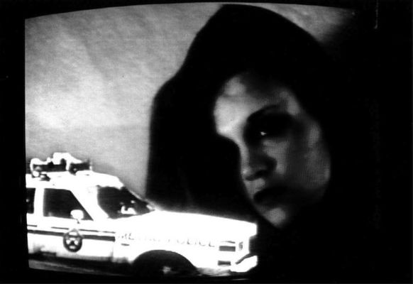 Grainy, black-and-white image of a police car overlayed on footage of a woman's face. Her face is largely in shadows.
