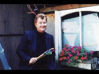 A person is in front of a wooden shed holding gardening tools. They are wearing a navy blue suit with a black turtleneck underneath with short dirty blonde hair. On the right hand side there is a window and a window sill flower box with purple and red flowers. They are smiling.