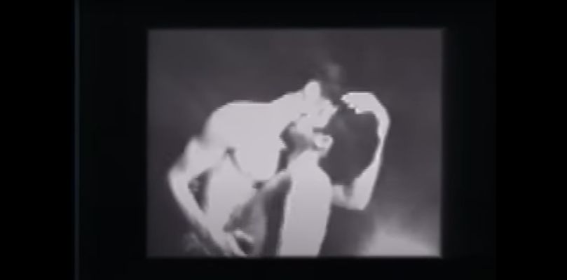 Two fair skinned men stand passionately entwined in a kiss while caressing each other. The setting appears to be a plain studio with a black background and a strong backlight shining at the camera. The still is in black and white.