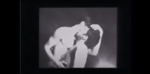 Two fair skinned men stand passionately entwined in a kiss while caressing each other. The setting appears to be a plain studio with a black background and a strong backlight shining at the camera. The still is in black and white.