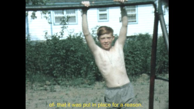 A young person with short orange hair is shirtless and in gray shorts holding onto a metal bar with their arms above their head. They are on dirt with bushes in the background. There is yellow text at the bottom that reads "of- that it was put in place for a reason."
