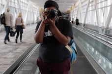 A person in a black t-shirt and maroon pants holds a camera. The person has dark brown skin and is looking into the camera to take a photo. They are standing on a mechanized walkway in an airport with various people walking back and behind the person.