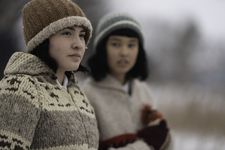 Two people with the one on the right out of focus and the one on the left in focus. The background is blurred but noticeably snowy outdoors. The person on the left is wearing a brown beanie. They have short dark brown hair. They are wearing a gray, beige, brown sweatshirt. The person on the right has short brown hair and is wearing a light blue and white beanie. They are also wearing a beige sweatshirt with a red line going over the torso. The one on the right is looking at the one on the left as the one on the left looks off into the distance.