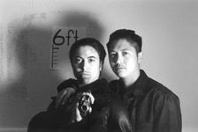 There is a black and white filter over the image. There are two people looking into the camera standing back to chest. The one on the left has their back against the chest of the one on the right. The right one has their hand on the right shoulder of the left one. The one on the left is holding a gun and pointing it at the camera wearing a black leather jacket with a furry collar. They have short slicked back hair and a goatee. The one on the right has short black hair with the sides faded and wearing a black jacket and a black t shirt underneath.