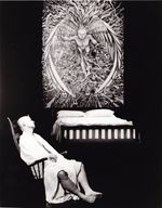Black and white image of a pale male figure in a dark bare room. The person is wearing nothing but a draped white sheet and is resting on a rocking chair with their eyes closed. Behind them there is a bed and a large angelic tapestry above the bed.