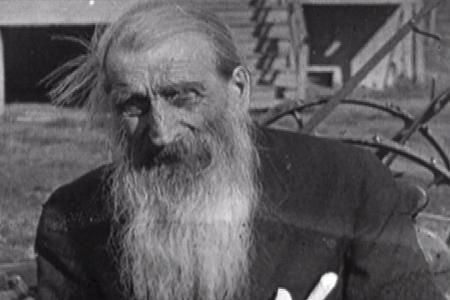 An archival black and white photograph of an elderly man in the the Canadian prairies, circa 1920-1940. The man has long light hair and a long light beard, and he wears a black coat. The stairs of a homestead are visible in the background.