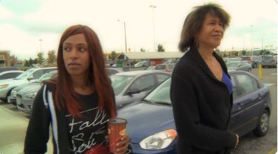 Two genderfluid people walk through a parking lot together, possibly a parent-child relationship. One appears older and is a light skinned Black person wearing a black blazer and blue shirt with a dark brown bob hairstyle, and the other is a younger dark skinned Black person wearing a black zipper hoodie and a dark graphic t-shirt, holding a coffee cup.