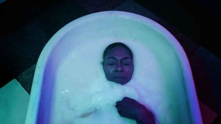 A person lays in a white tub with only their face and right hand sticking out of the white goo they are lying in. They have dark brown skin with their eyes closed and they have a buzz cut.