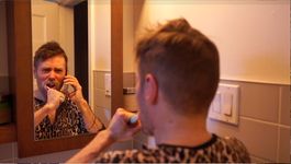 A person is squinting into a mirror while on the phone and brushing their teeth. They are wearing a cheetah print t-shirt and they have short light brown hair with facial stubble.