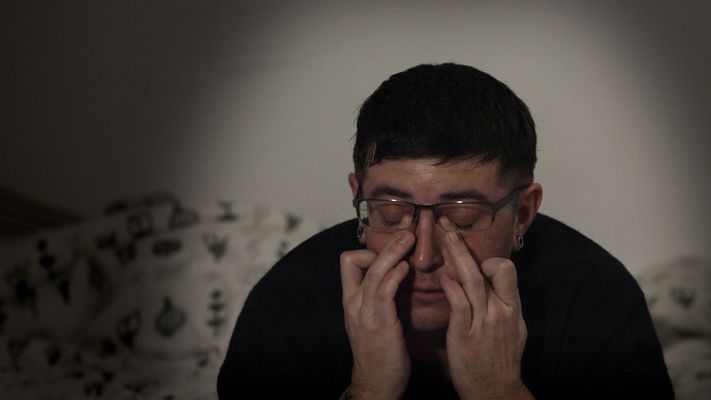 A person sitting hunched over rubbing their tear ducts with their middle fingers. The person has light skin, short black hair, glasses and a black shirt on. The background is a couch with a white blanket with black designs all over it.