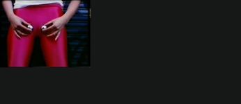 Closeup shot of a white person's groin area where they are holding two metal balls near their pubic area to represent testicles. They are wearing bright pink leggings and a white short sleeve shirt. They stand in an alleyway in front of a black garage door with graffiti.