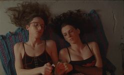 Two people are lying next to each other on a blanket, each with their hair splayed out loosely onto the floor above their head. The person on the left has long light brown hair, is wearing clear glasses, and wears a black-and-red tank top. The person on the right has long brown hair, a black-and-red tank top with a different design, and fishnet-arm sleeves. The person on the left is lighting an object with a lighter that they are holding in their right hand. The person on the right has their arm laid out across their chest, reaching gently towards the object being lit. They are both looking at the object as it gets lit.