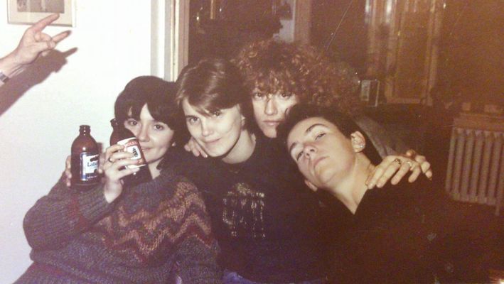Four people (Holly Dale, Shaggie, Janis Cole, and Minx), pose together in celebration of Shaggie’s release from the Prison for Women. The leftmost person is holding a beer up near their face and wearing a patterned sweatshirt with light skin and short brown hair. The next person has light skin, short brown light hair and a patterned sweater with their left arm around the person to their left holding a beer in their hand as well. The person to the right of them has long curly light brown hair. Their head is perched between the person to their left and right so you can't see their body. The last person on the far right is leaning their head over. They have light skin, short black hair parted in the middle, and wear a black top.