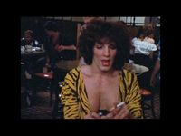 A person looking at something in their left palm while mid sentence. They are holding a cigarette in their right hand. They have light skin and short curly dark brown hair. They have a gold cross necklace around their neck, wearing a yellow-and-black animal print top with a deep v-neck. The background is a crowded restaurant with a bunch of people sitting at various tables and waiters in motion.