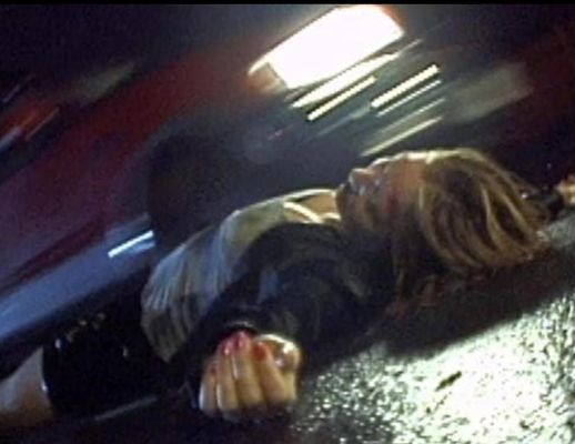 A person is lying down in the road as a red car rushes by them. The street looks wet as if it rained. The person is lying sprawled out with blonde hair, a gray t-shirt and black leather skirt and a black jacket. They have their nails painted pink and their forehead seems to be bloodied.