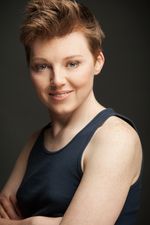 Studio headshot of Amy Fox, a white transgender woman with tousled short brown hair. Amy wears a navy tank top, and stares down at the camera with a devious grin.