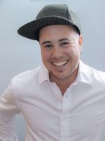 Professional portrait of Rémy Huberdeau, a white Franco-Manitoban trans man, wearing a white button-up shirt and a snapback hat. They stand and smile confidently with their hands on their hips in front of a light studio background.