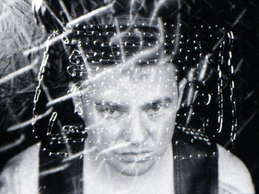 A creative self-portrait taken by Malic Amalya. Malic Amalya has fair skin, short dark hair, and is wearing a black tank top. They look towards the camera pointed above with their eyes only, posing with a serious and focused expression. The image is a black-and-white photograph with a surreal and intense atmosphere created by eerie lighting, the dark background, superimposed metallic chain elements and abstract fragmented patterns overtop the portrait.