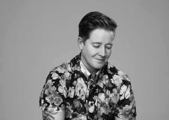 A greyscale studio portrait of Ivan Coyote, a non-binary person with light skin and short dark hair, captured in a candid calm moment. They are wearing a floral-patterned button-up shirt and are looking downwards with a gentle smile.