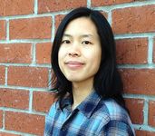 Bo Rittapa standing in front of a red brick wall. He has light skin, shoulder-length dark hair, and is wearing a blue plaid shirt. He has a calm and relaxed expression while slightly smiling at the camera.