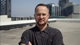 Picture of Finn Paul taken on a rooftop in a cityscape with clear daytime skies. He gives a squinty, focused smile illuminated by strong rays of sunshine. They wear a short-sleeved dark pattern button up shirt accessorized with clear framed glasses, a small earring and dark trimmed facial hair.