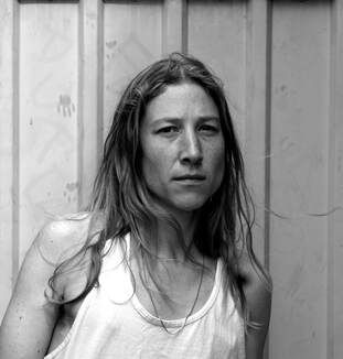 Black-and-white photograph of Wren Warner with a composed expression standing in front of a metal facade resembling a shipping container. They are wearing a white sleevelss tank top and their body is in a relaxed, candid posture which contrasts the serious and focused look in their eyes.