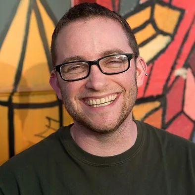 Portrait of Sam Berliner with a beaming smile on a sunny day in front of colourful street art. He has fair skin, black-rimmed rectangular glasses and short brown hair, and is wearing a dark green shirt.
